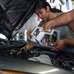 A woman watches as a man pours STP synthetic motor oil to show her how to change the car's oil.