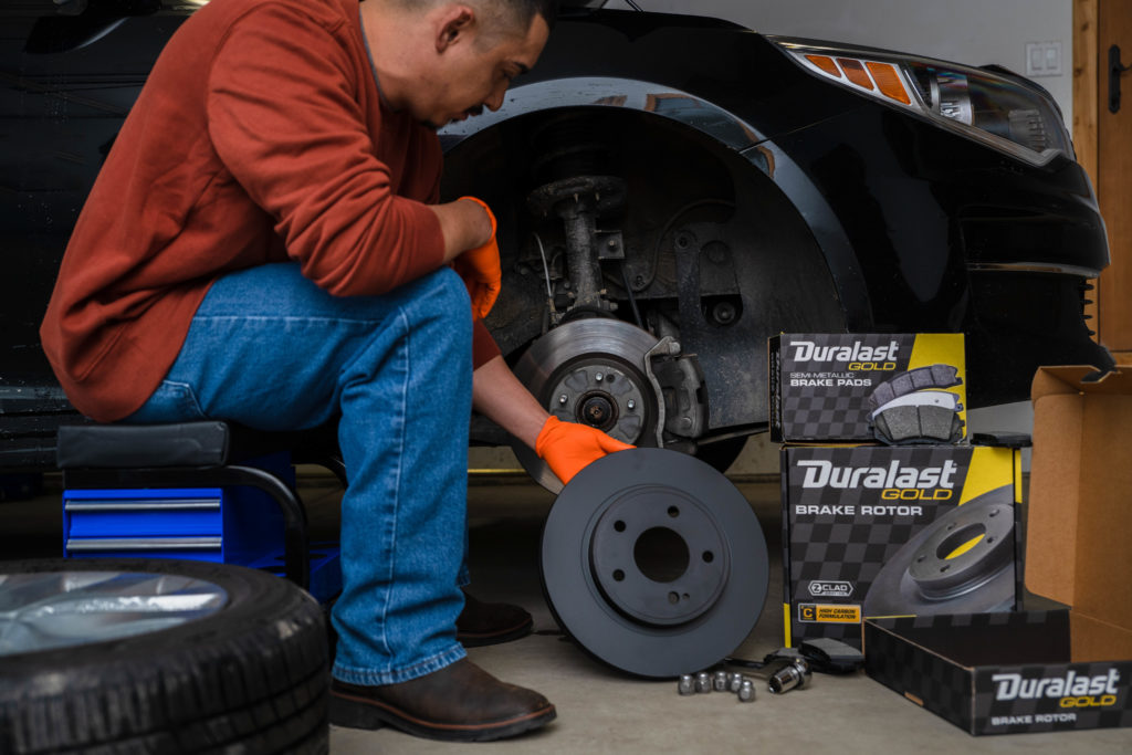 Duralast gold pads rotor boxes rotor in hand in front of car with removed wheel